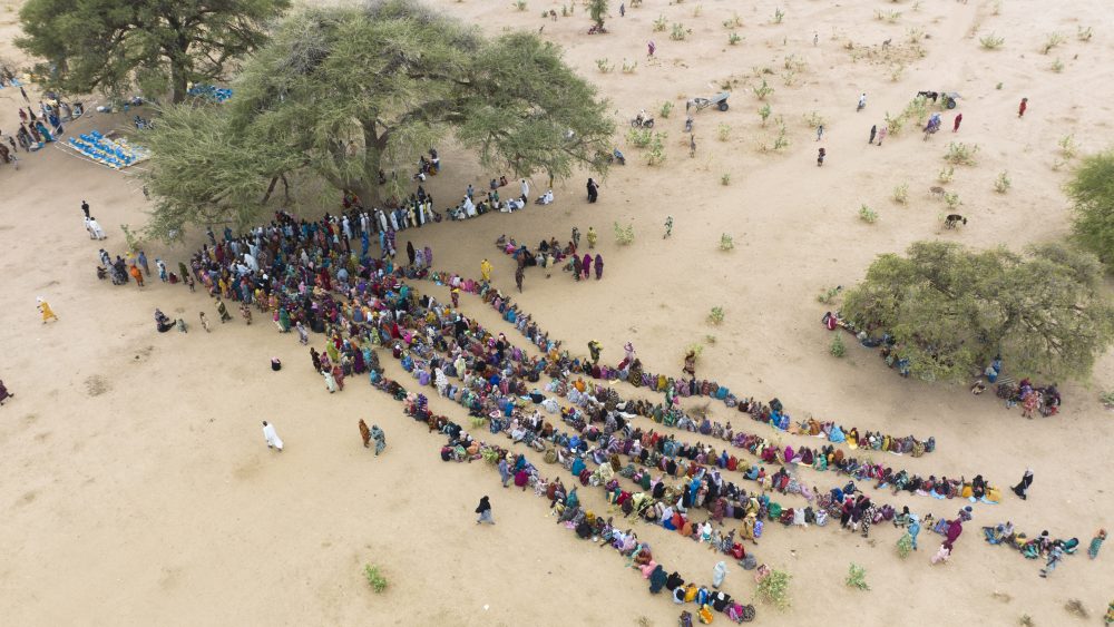 Aerial view of hundreds of people seated in long curved lines under shady trees in a dry, sandy landscape.
