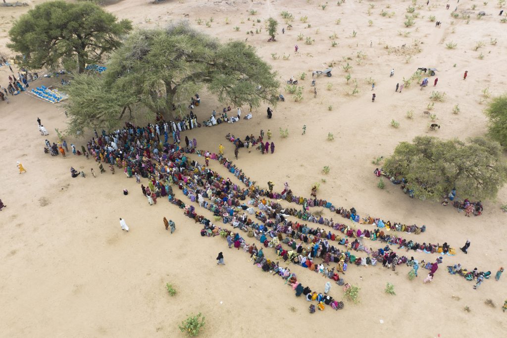 Aerial view of hundreds of people seated in long curved lines under shady trees in a dry, sandy landscape.