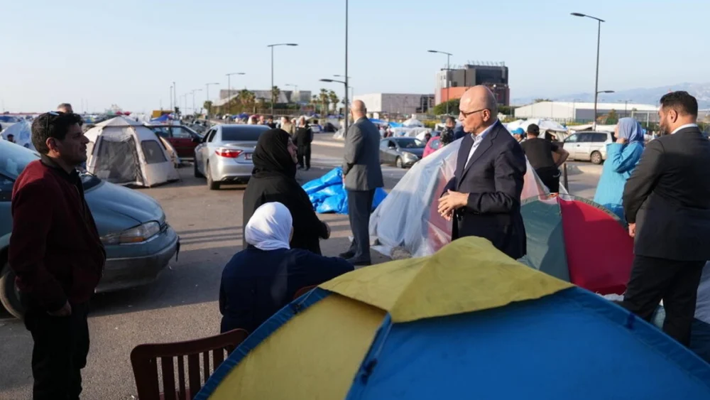 Temporary refugee camp set up in a parking lot with colorful tents, cars, and people talking nearby on a sunny day, urban backdrop nearby