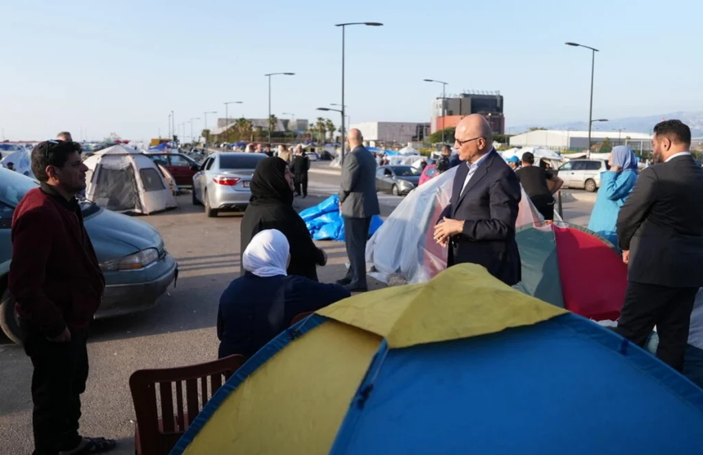 Temporary refugee camp set up in a parking lot with colorful tents, cars, and people talking nearby on a sunny day, urban backdrop nearby