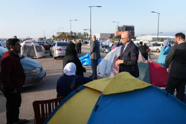 Temporary refugee camp set up in a parking lot with colorful tents, cars, and people talking nearby on a sunny day, urban backdrop nearby