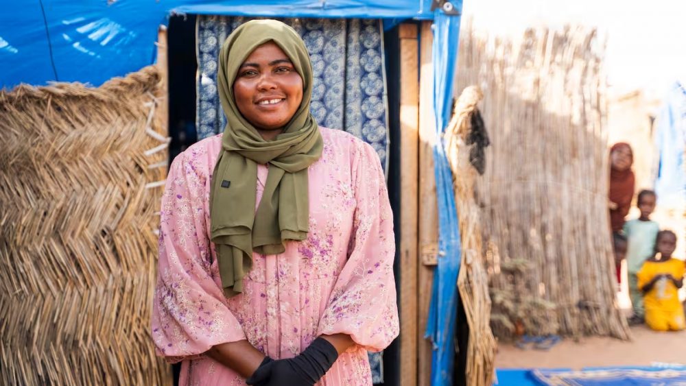 Smiling woman in a green hijab and pink floral dress stands at the doorway of a blue tent in a straw-walled camp, with children in the background.