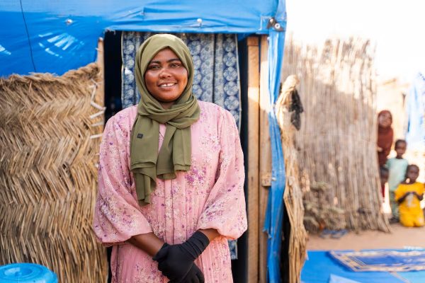 Smiling woman in a green hijab and pink floral dress stands at the doorway of a blue tent in a straw-walled camp, with children in the background.