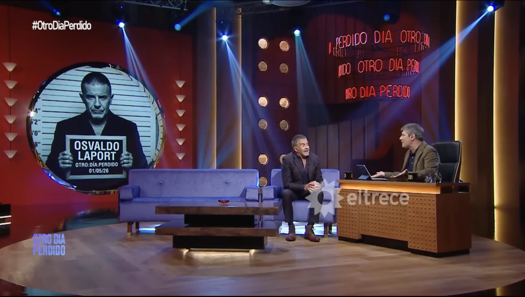 Large TV studio set with a circular mugshot portrait of Osvaldo Laport on the left, showing the date 01/05/26 and 'OTRO DIA PERDIDO'; two men sit and chat on blue couches, host desk on the right, 'eltrece' logo visible.