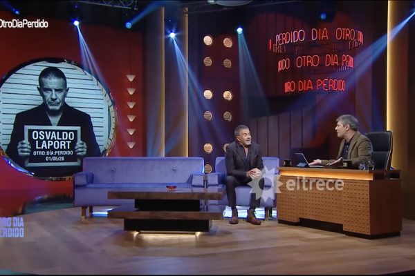 Large TV studio set with a circular mugshot portrait of Osvaldo Laport on the left, showing the date 01/05/26 and 'OTRO DIA PERDIDO'; two men sit and chat on blue couches, host desk on the right, 'eltrece' logo visible.