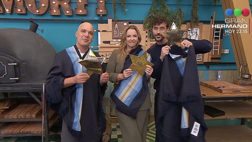 Four graduates in dark blue robes and one person in olive attire pose together, each holding gold star awards in a craft workshop with a wood-fired oven and tools in the background, Gran Hermano logo visible in the top-right.