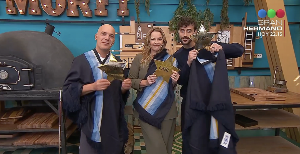 Four graduates in dark blue robes and one person in olive attire pose together, each holding gold star awards in a craft workshop with a wood-fired oven and tools in the background, Gran Hermano logo visible in the top-right.