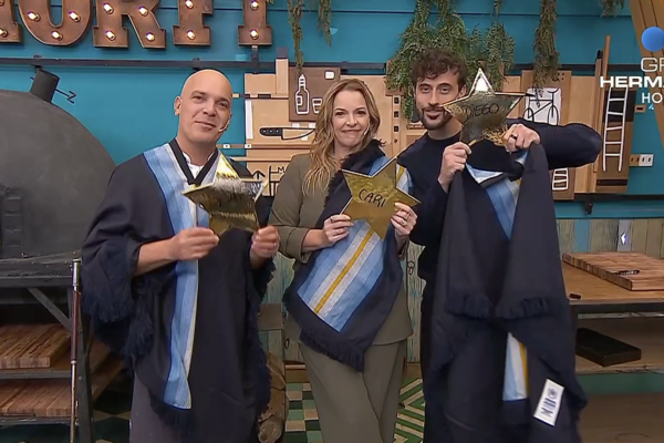 Four graduates in dark blue robes and one person in olive attire pose together, each holding gold star awards in a craft workshop with a wood-fired oven and tools in the background, Gran Hermano logo visible in the top-right.