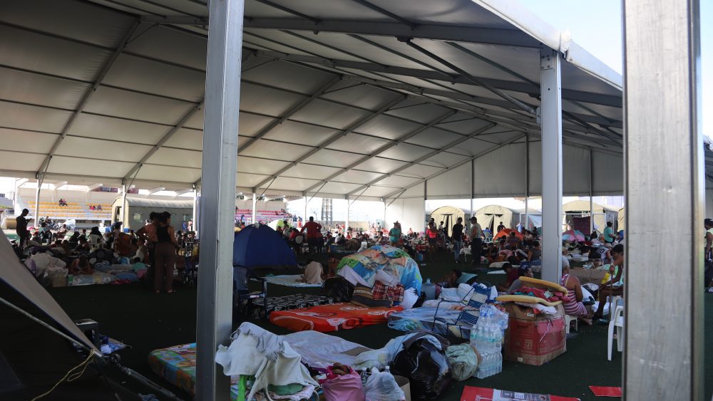 Crowded relief camp under a large metal-framed tent with many people sitting on mats and blankets amid scattered belongings and tents outside.