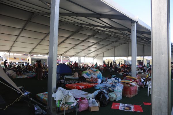 Crowded relief camp under a large metal-framed tent with many people sitting on mats and blankets amid scattered belongings and tents outside.