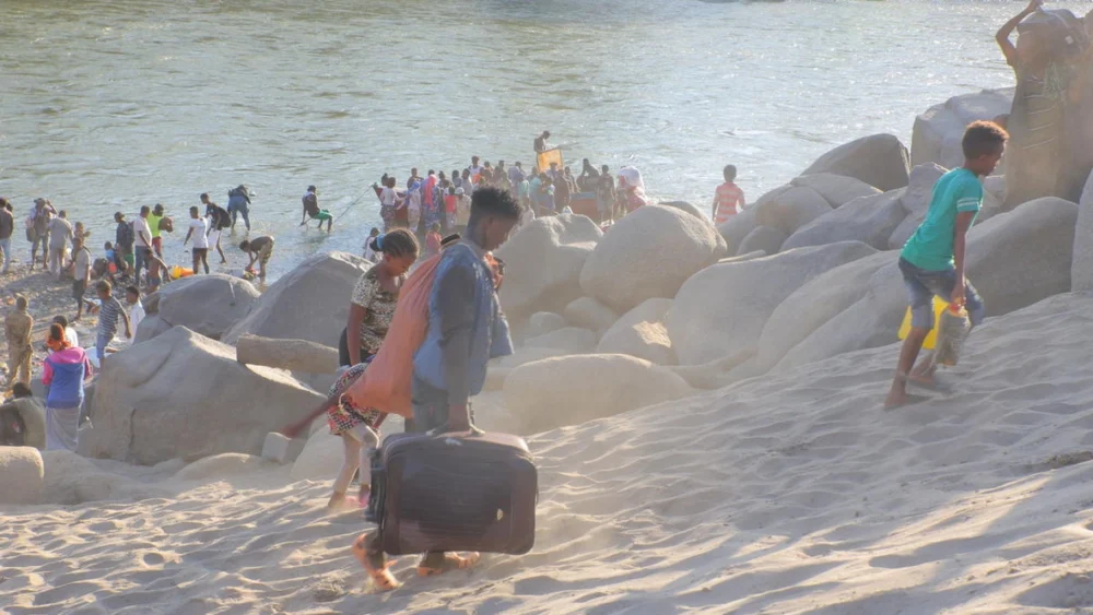 Crowded rocky beach by the water; people climb on large boulders while some carry bags and luggage on the sand.
