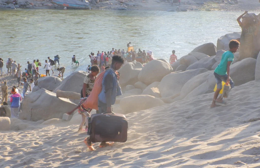 Crowded rocky beach by the water; people climb on large boulders while some carry bags and luggage on the sand.