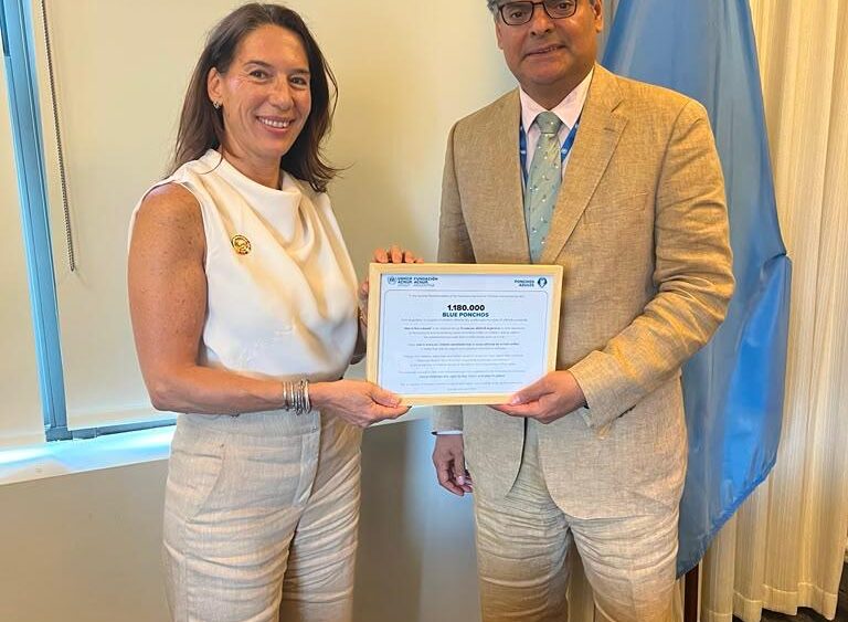 Two professionals stand side by side, smiling, as they hold a framed certificate together in a formal office setting with a blue flag in the background.