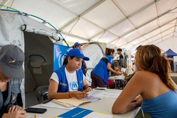 Volunteer in a blue cap and vest sits at a table inside a large tent, helping a woman fill forms.
