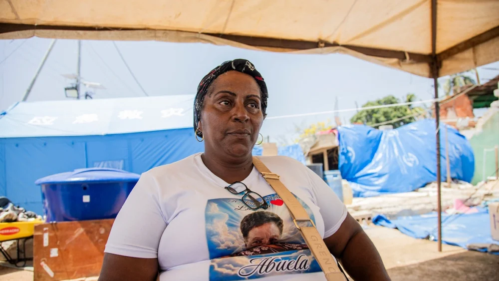 Woman sitting under a canopy at an outdoor relief site, wearing a white T-shirt with a graphic that includes the word 'Abuela' and a crossbody bag strap visible.