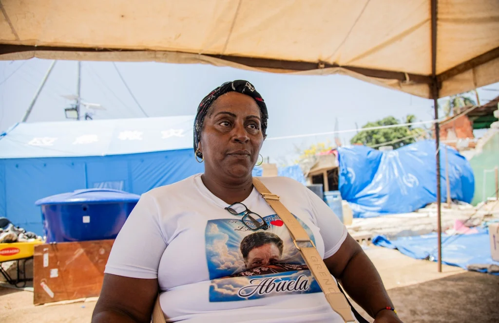 Woman sitting under a canopy at an outdoor relief site, wearing a white T-shirt with a graphic that includes the word 'Abuela' and a crossbody bag strap visible.