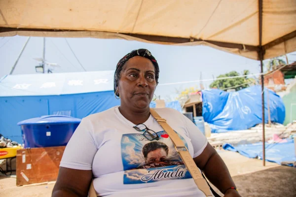Woman sitting under a canopy at an outdoor relief site, wearing a white T-shirt with a graphic that includes the word 'Abuela' and a crossbody bag strap visible.