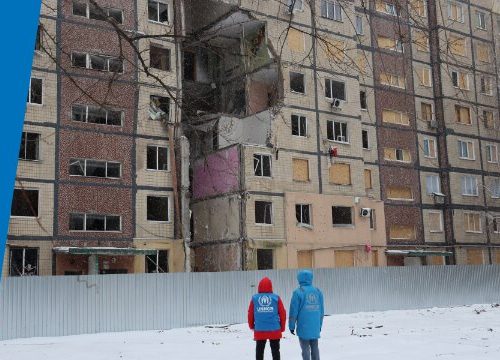 Dos trabajadores de ACNUR caminan en la nieve frente a un edificio dañado
